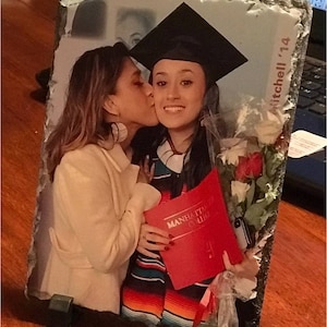 May include: A black and white photo of two women, one wearing a graduation cap and gown, the other wearing a white jacket. The woman in the graduation gown is holding a red book that says "Manhattan College". The photo is on a slate plaque with the text "Mitchell '14" written on it.