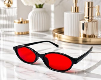 May include: Oval-shaped sunglasses featuring a glossy black plastic frame and vibrant red lenses. The eyewear is positioned on a white marble surface, showcasing a sleek, minimalist design with thin temples and a smooth, continuous bridge structure.