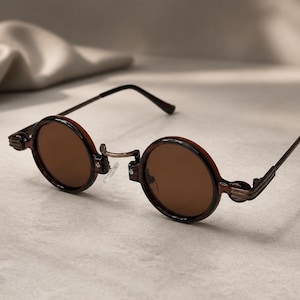May include: Round sunglasses featuring dark brown lenses and a metallic bronze frame. The circular frames have a distinct ridged detail at the hinges, connecting to thin, straight metal temples. The bridge is curved with clear nose pads.