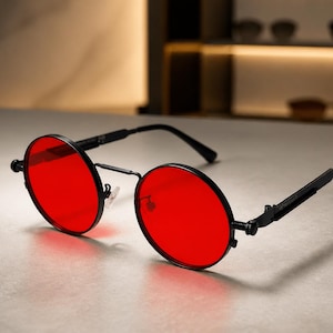 May include: Round sunglasses featuring vibrant red lenses and a thin black metal frame. The eyewear includes adjustable nose pads and slender black temples with a textured, mechanical-style hinge detail at the connection point to the frame.