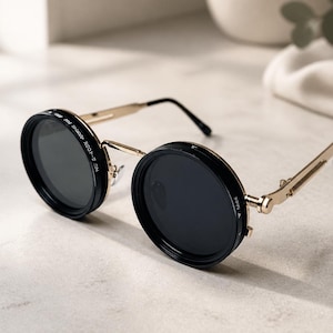 Peut inclure: Lunettes de soleil rondes avec des verres circulaires noir foncé entourés de montures noires épaisses et de branches en métal doré. Le côté de la monture porte l'inscription blanche WC-400X. Les branches présentent une charnière mécanique.