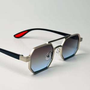 Peut inclure: Lunettes de soleil à monture argentée de forme octogonale avec verres bleus dégradés. Les branches sont noires avec des pointes rouges. Le pont est une fine barre argentée.