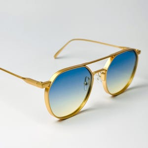 May include: Gold-framed sunglasses with blue and yellow gradient lenses. The aviator-style frames have a double bridge and a sleek, modern design. The lenses transition from blue to yellow.