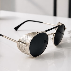 May include: Round sunglasses with dark lenses, a thin silver-tone metal frame, and a double bridge design. The sides feature perforated metal side shields, and the temples are finished with black plastic tips for a secure fit over the ears.