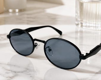 May include: Oval sunglasses featuring a thin black metal frame and dark gray tinted lenses. The eyewear has slender temples and adjustable nose pads, providing a sleek and simple design with a classic rounded shape for everyday eye protection.