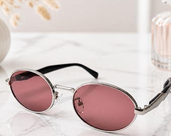 May include: Oval-shaped sunglasses featuring a polished silver metal frame and tinted rose-colored lenses. The temples are black, and the bridge includes adjustable nose pads. These glasses have a classic, rounded silhouette with a metallic finish.