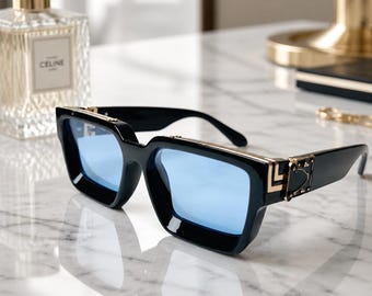 May include: Black sunglasses featuring thick frames and light blue lenses. The temples are accented with gold-toned metal hardware, including a geometric design near the hinges and small studs along the top edge of the frame front.