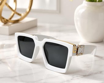 May include: White sunglasses featuring dark gray lenses and a thick, square-shaped frame. The temples are accented with gold-toned metal hardware, including a geometric detail near the hinge and a small, rectangular plate with a metallic finish.