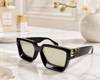 May include: Black sunglasses with thick frames and light-tinted lenses. The temples feature a gold-tone metal accent with a geometric design and small studs. The bridge is straight, and the overall construction is bold with a glossy finish.