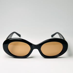 May include: Black oval sunglasses with brown lenses. The sunglasses have a thick, glossy black frame and a classic, rounded shape. The lenses are a warm, light brown color, offering a contrast to the dark frame. The arms are also black.