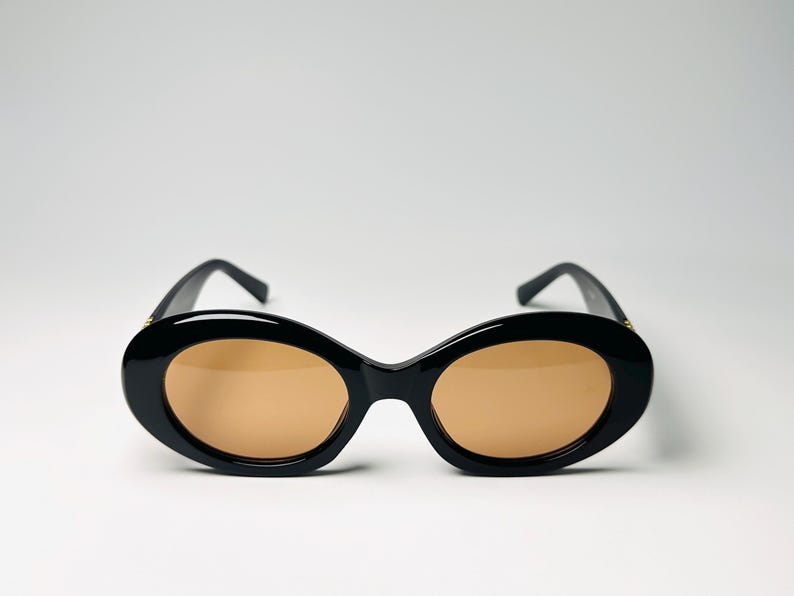 May include: Black oval sunglasses with brown lenses. The sunglasses have a thick, glossy black frame and a classic, rounded shape. The lenses are a warm, light brown color, offering a contrast to the dark frame. The arms are also black.