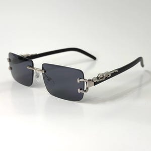 May include: Rimless sunglasses with dark gray lenses and black temples. The silver-toned metal bridge and hinges feature a decorative jaguar design. The sunglasses have a rectangular shape and are set against a white background.
