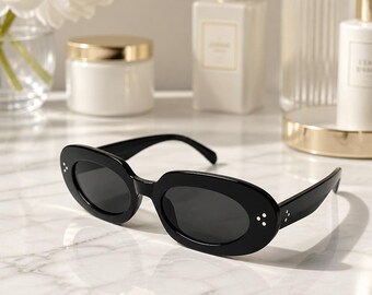 May include: Black oval-shaped sunglasses featuring dark tinted lenses and a glossy frame. Each side of the front frame is accented with three small, vertically aligned silver-toned dots, providing a subtle detail to the solid black plastic construction.