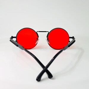 May include: Round sunglasses featuring vibrant red lenses and a thin black metal frame. The temples are black and cross over each other at the bottom, showcasing a minimalist design with a bridge connecting the two circular lens rims.