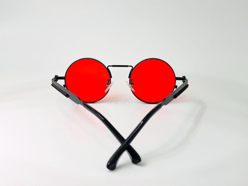 May include: Round sunglasses featuring vibrant red lenses and a thin black metal frame. The temples are black and cross over each other at the bottom, showcasing a minimalist design with a bridge connecting the two circular lens rims.