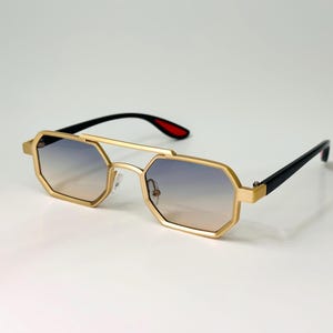 May include: Gold-framed sunglasses with a unique octagonal shape and gradient lenses. The arms are black with red accents near the hinges. The sunglasses have a modern, stylish design.