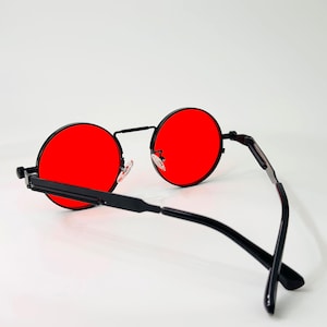 May include: Round sunglasses featuring vibrant red lenses and a thin black metal frame. The design includes a straight bridge, adjustable nose pads, and slender black temples with a textured detail along the side of the frame arms.