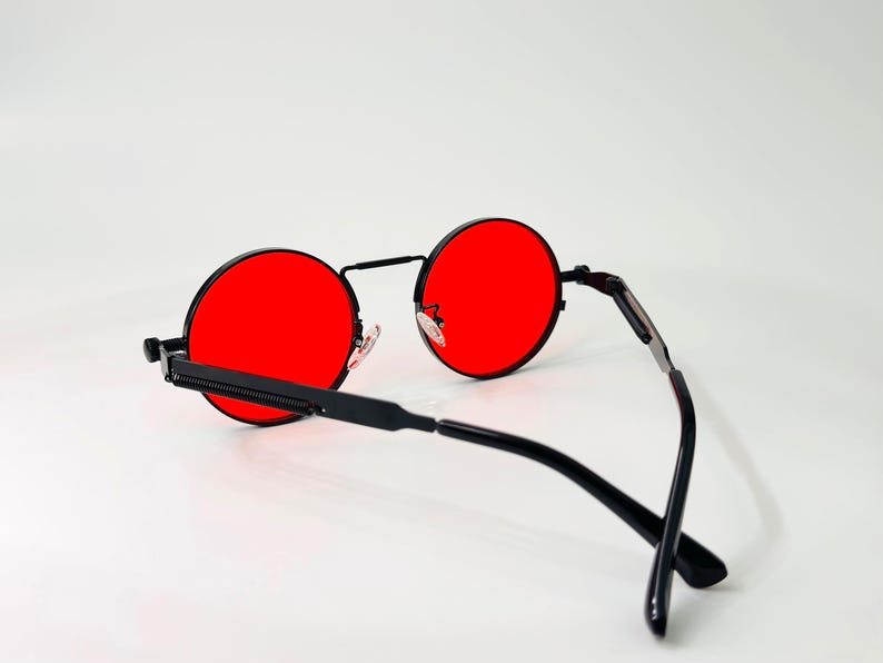 May include: Round sunglasses featuring vibrant red lenses and a thin black metal frame. The design includes a straight bridge, adjustable nose pads, and slender black temples with a textured detail along the side of the frame arms.