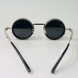 May include: Circular black sunglasses with silver frames and black arms. The lenses are dark, and the frames have a vintage style. The sunglasses are set against a plain white background.