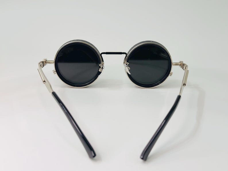 May include: Circular black sunglasses with silver frames and black arms. The lenses are dark, and the frames have a vintage style. The sunglasses are set against a plain white background.