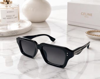 May include: Black sunglasses with a thick rectangular frame and dark tinted lenses. The temples feature silver-toned metal accents and three small circular studs on each side of the front frame, providing a structured and bold appearance.