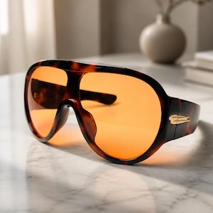 May include: Oversized shield sunglasses featuring a dark brown tortoiseshell frame and vibrant orange tinted lenses. The frame has a continuous wrap-around design with a gold-toned metal accent on the temple hinge, providing a bold look.