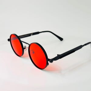 May include: Round sunglasses featuring vibrant red lenses and a matte black metal frame. The eyewear includes thin, straight temples with a mechanical hinge design, a thin bridge, and adjustable nose pads, providing a distinct circular silhouette.
