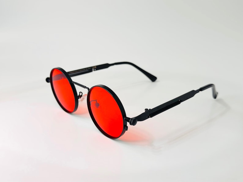 May include: Round sunglasses featuring vibrant red lenses and a matte black metal frame. The eyewear includes thin, straight temples with a mechanical hinge design, a thin bridge, and adjustable nose pads, providing a distinct circular silhouette.