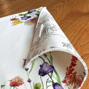 K&ouml;nnte beinhalten: Wei&szlig;e Tischsets aus Baumwolle mit einem Blumendesign mit violetten, roten und gelben Blumen. Das Tischset hat einen wei&szlig;en Rand und ein wei&szlig;es Etikett, das an der Ecke befestigt ist.