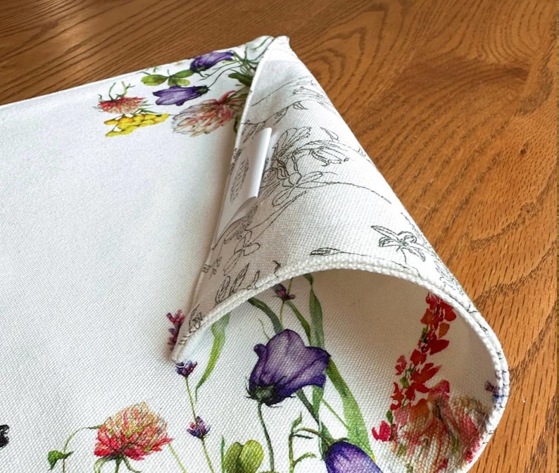 K&ouml;nnte beinhalten: Wei&szlig;e Tischsets aus Baumwolle mit einem Blumendesign mit violetten, roten und gelben Blumen. Das Tischset hat einen wei&szlig;en Rand und ein wei&szlig;es Etikett, das an der Ecke befestigt ist.