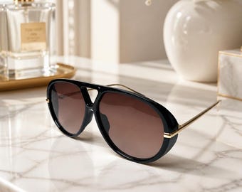 May include: Black aviator sunglasses featuring a thick, bold frame with a double bridge design and thin gold-tone metal temples. The lenses are a gradient brown tint, providing a classic look with a modern, structured silhouette and clean lines.