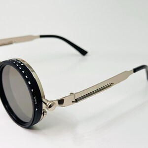 May include: Close-up of steampunk-style sunglasses with round, grey lenses. The frames are silver with black accents and hinges. The temples have a spring design. The sunglasses are on a white surface.