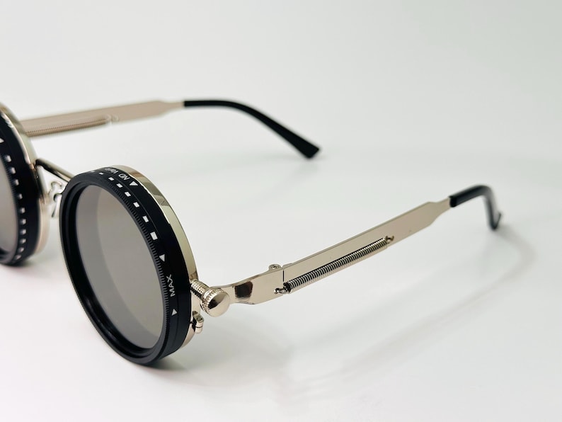 May include: Close-up of steampunk-style sunglasses with round, grey lenses. The frames are silver with black accents and hinges. The temples have a spring design. The sunglasses are on a white surface.