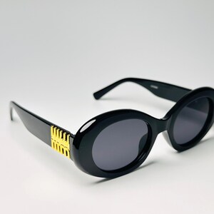 May include: Black oval sunglasses with dark gray lenses. The frames are thick and glossy, with gold-colored accents on the temples. The sunglasses have a classic, retro style.