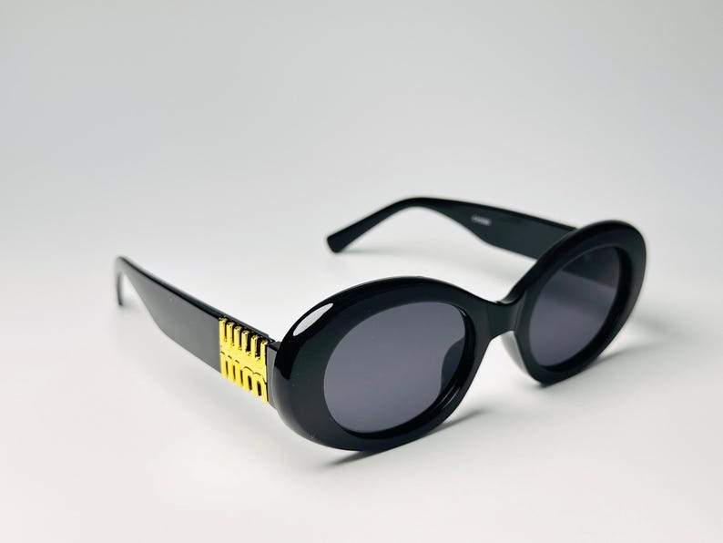 May include: Black oval sunglasses with dark gray lenses. The frames are thick and glossy, with gold-colored accents on the temples. The sunglasses have a classic, retro style.