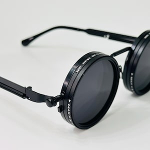 May include: Black round sunglasses with dark lenses. The sunglasses have a unique design with a spring hinge and a thin, rectangular arm. The lenses are surrounded by a black frame with text. The sunglasses are set against a white background.
