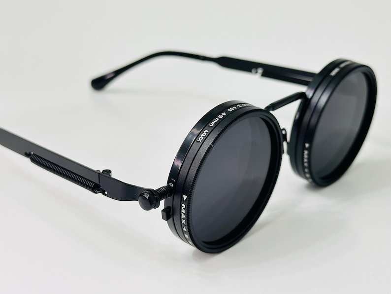 May include: Black round sunglasses with dark lenses. The sunglasses have a unique design with a spring hinge and a thin, rectangular arm. The lenses are surrounded by a black frame with text. The sunglasses are set against a white background.