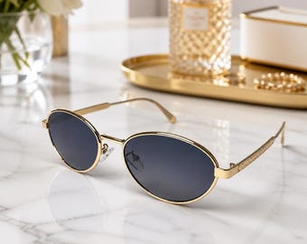 May include: Oval sunglasses featuring a thin gold-tone metal frame and dark gray lenses. The temples have a textured pattern along the sides, and the bridge includes adjustable nose pads. These glasses are designed with a classic silhouette.