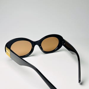 May include: Black oval sunglasses with brown lenses. The sunglasses have a gold accent on the temples. The frames are thick and the lenses are a light brown color. The sunglasses are angled slightly, showing the side of the frame.