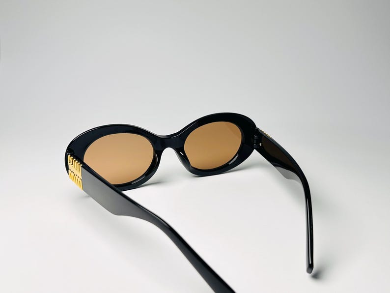 May include: Black oval sunglasses with brown lenses. The sunglasses have a gold accent on the temples. The frames are thick and the lenses are a light brown color. The sunglasses are angled slightly, showing the side of the frame.