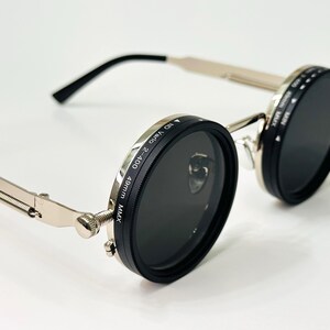 May include: Round sunglasses with black lenses and silver frames. The lenses are surrounded by black rings with white text. The arms are silver with black ear pieces. The sunglasses are on a white surface.