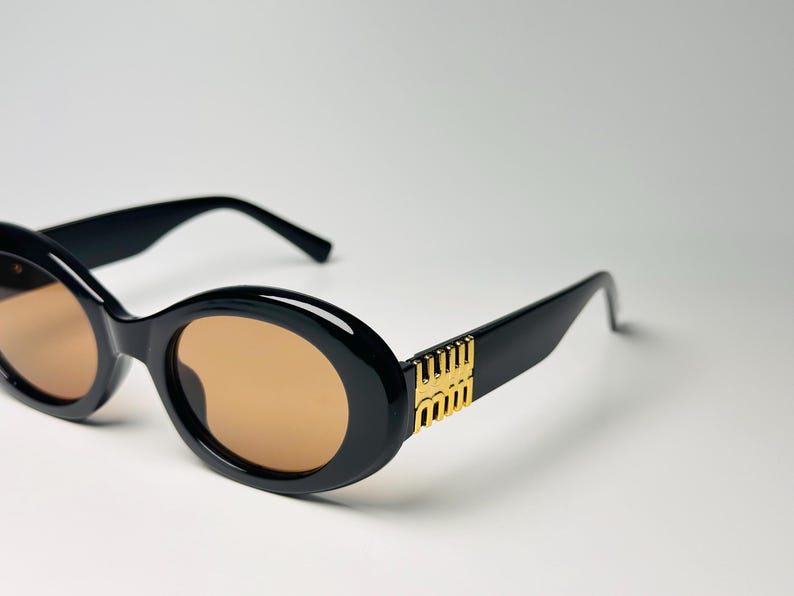 May include: Black oval sunglasses with brown lenses. The frames have a thick, glossy finish. Gold-colored accents are on the temples. The sunglasses are set against a white background.