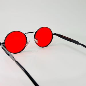 May include: Round sunglasses featuring vibrant red lenses and a thin black metal frame. The eyewear includes adjustable nose pads and slender black temples, with visible text printed on the inner side of the arm indicating the design origin.
