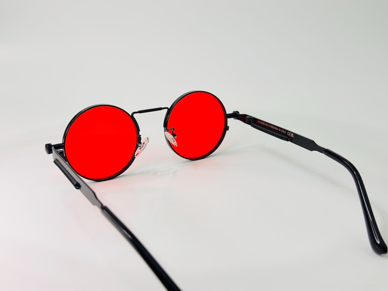 May include: Round sunglasses featuring vibrant red lenses and a thin black metal frame. The eyewear includes adjustable nose pads and slender black temples, with visible text printed on the inner side of the arm indicating the design origin.