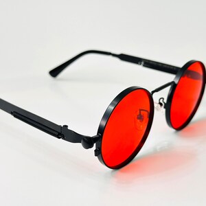 May include: Round sunglasses featuring vibrant red lenses and a matte black metal frame. The temples are designed with a distinct coiled spring detail near the hinges, and the bridge includes adjustable nose pads for a secure fit.