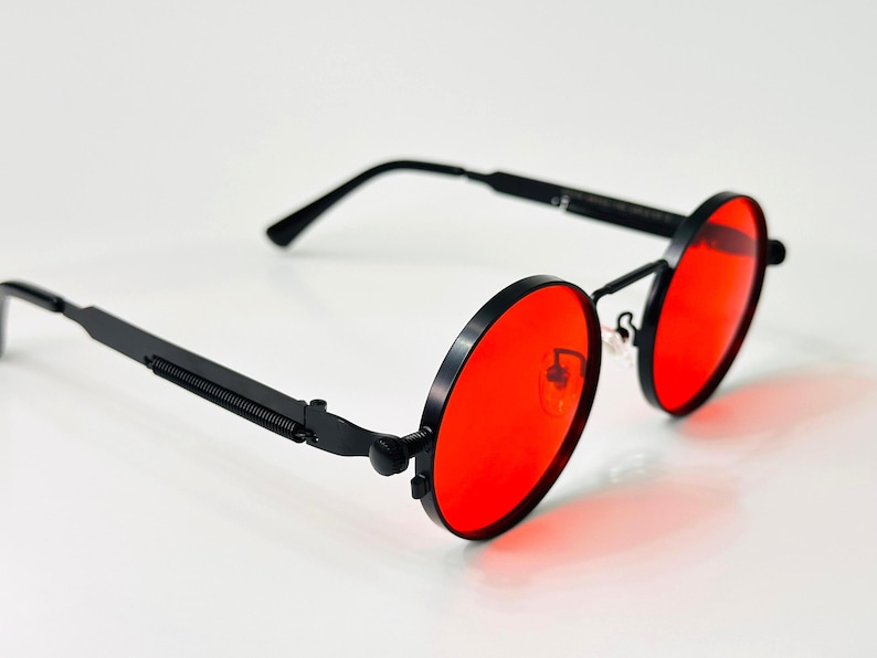 May include: Round sunglasses featuring vibrant red lenses and a matte black metal frame. The temples are designed with a distinct coiled spring detail near the hinges, and the bridge includes adjustable nose pads for a secure fit.