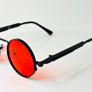 May include: Round sunglasses featuring vibrant red lenses and a matte black metal frame. The design includes thin, straight temples with a distinct hinge mechanism and adjustable nose pads, providing a clear view of the circular lens shape.
