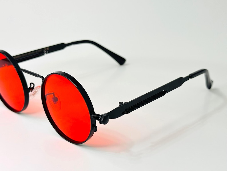 May include: Round sunglasses featuring vibrant red lenses and a matte black metal frame. The design includes thin, straight temples with a distinct hinge mechanism and adjustable nose pads, providing a clear view of the circular lens shape.