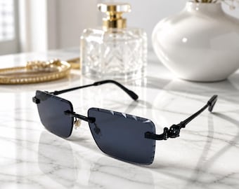 May include: Rimless sunglasses featuring rectangular dark gray lenses with a subtle etched pattern on the outer edges. The frames are black with thin metal temples and a decorative emblem detail near the hinges, providing a sleek look.