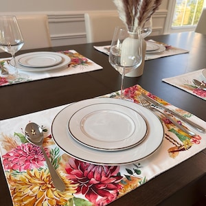 K&ouml;nnte beinhalten: Ein wei&szlig;er Platzteller mit einem Blumendesign mit rosa, gelben und violetten Blumen. Der Platzteller ist mit einem wei&szlig;en Teller, einer silbernen Gabel und einem Messer sowie einem silbernen L&ouml;ffel gedeckt.